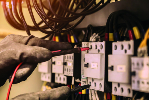 Manos enguantadas de un técnico usando un multímetro para probar un panel eléctrico con cables multicolores en un entorno industrial.